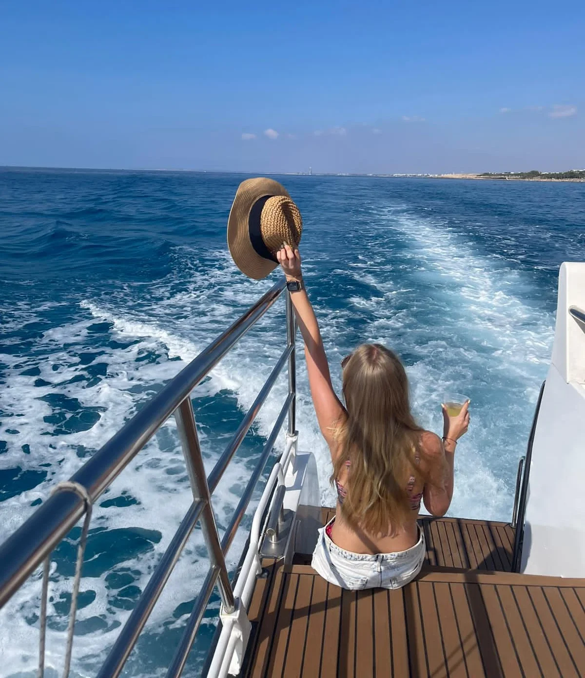 Guest holding a sun hat and a drink at the back of the catamaran, watching the Cape Greco coastline
