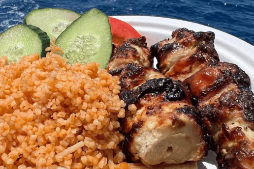 Plate of grilled chicken souvlaki with pourgouri, cucumber and tomato, with the sea in the background