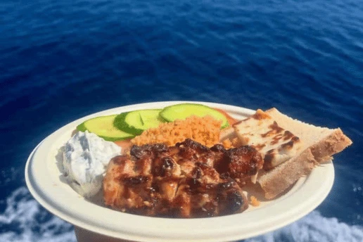 Plate of grilled chicken souvlaki, halloumi, pourgouri, tzatziki, bread and cucumber held over the sea
