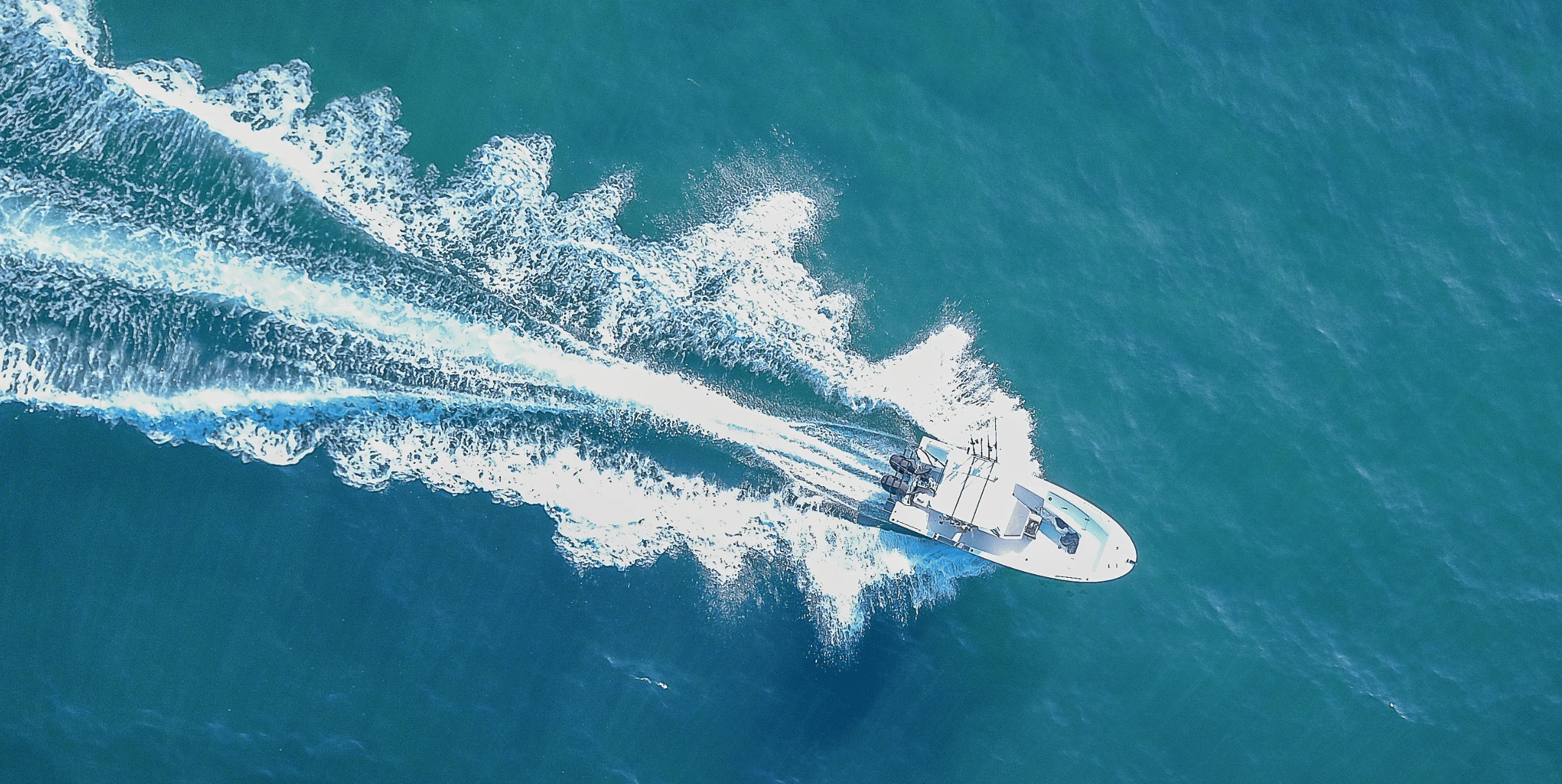 Aerial view of a white speedboat with twin outboard engines leaving a long white wake across blue-green sea