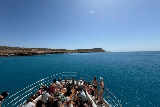 Guests crowding the bow of a boat as it heads across calm blue water towards the headland at Cape Greco