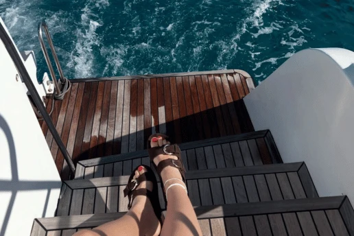 Feet in sandals on the teak swim steps at the stern of a catamaran, with the sea and wake behind