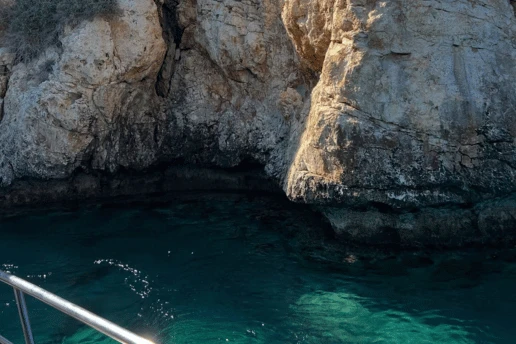 Sunlit limestone cliff and shaded cave over clear turquoise water, with a boat railing in the foreground
