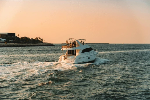 White flybridge motor yacht cruising at golden hour past a low coastline with palm trees