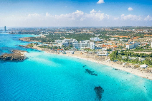 Aerial view of Nissi Beach in Ayia Napa, with a sandy shoreline, hotels and a small rocky islet in turquoise water