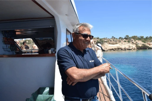 Crew member in a navy polo shirt and sunglasses standing at the deck rail, with a rocky coastline behind