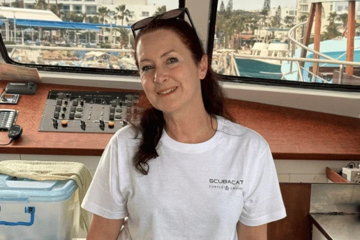 Smiling crew member in a white Scubacat Turtle Cruise T-shirt in the wheelhouse, with a harbour visible through the windows