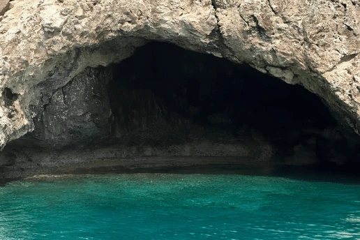 Dark sea cave entrance at water level, seen from a boat on calm turquoise water near Cape Greco