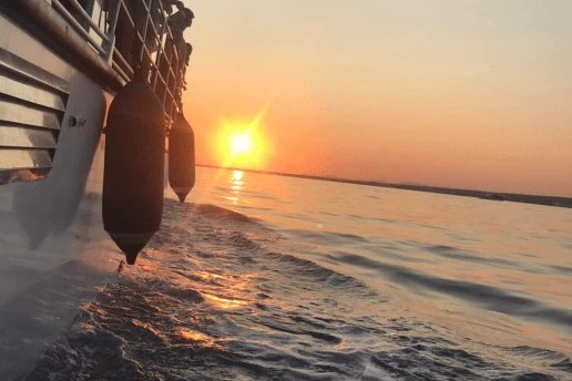 Sun setting over the sea, seen from the side of a moving boat with fenders hanging over the water