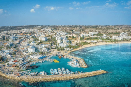 Aerial view of Ayia Napa harbour, with boats moored inside the breakwater, the town behind and a sandy beach to the right
