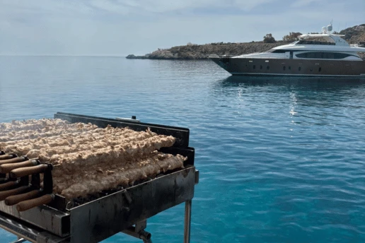 Charcoal grill loaded with chicken souvlaki skewers on a boat, with a large motor yacht anchored in the bay behind