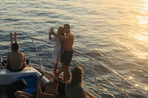 Guests on the bow of a boat at golden hour, a couple taking a selfie by the rail and others sitting on deck