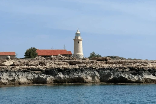 Cape Greco lighthouse and red-roofed buildings on a low rocky headland, seen from the sea under a blue sky