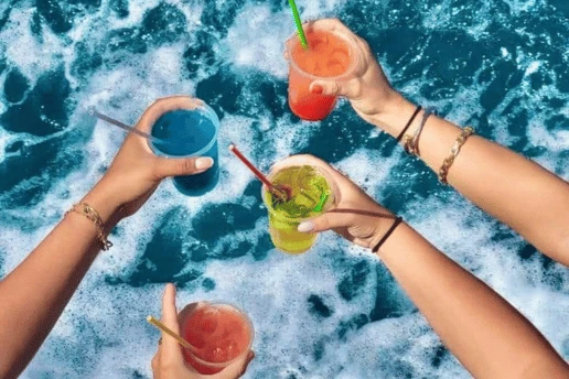 Four hands holding colourful drinks with straws above the white wake behind a boat