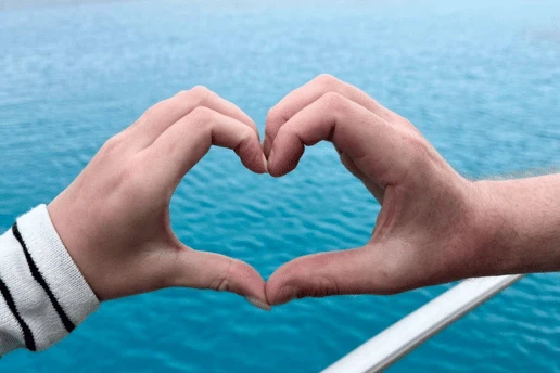 Two people's hands forming a heart over turquoise water, with the boat's rail below and a rocky headland behind