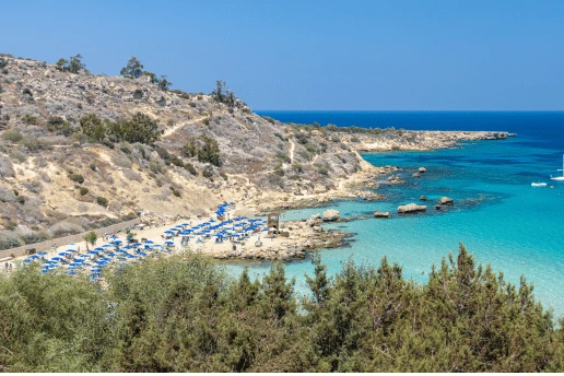 Konnos Bay seen from the hillside, with blue parasols on a sandy cove, pine trees and boats in turquoise water