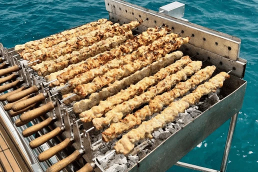 Charcoal grill with rows of chicken souvlaki skewers mounted at the stern of a boat, over open sea