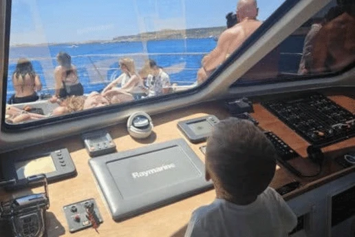 Young child looking out from the wheelhouse over the helm console, with guests relaxing on the foredeck and the sea beyond