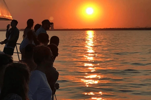 Silhouetted guests lined along a boat rail, watching the sun set over the sea with a distant coastline behind