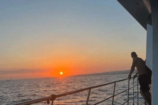 Guest leaning on the side rail of a boat at sunset, with the sun on the horizon and a distant coastline