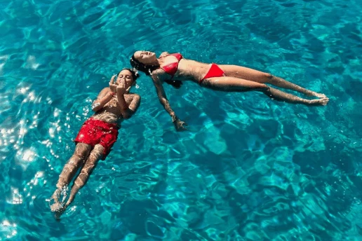 Two swimmers floating on their backs in clear turquoise water, seen from the deck above